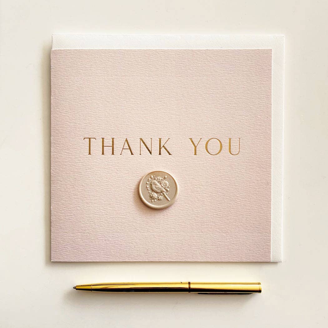 A square blush pink thank-you card with gold foil text reading 'THANK YOU' and a small gold seal, shown with a white envelope behind it and a gold pen below on a light neutral background.
