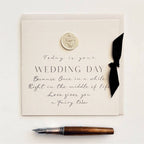 A square cream-colored wedding card with elegant black script and serif text reading 'WEDDING DAY' and a decorative quote, styled with a small round wax seal, a black ribbon bow, and a fountain pen on a white background.