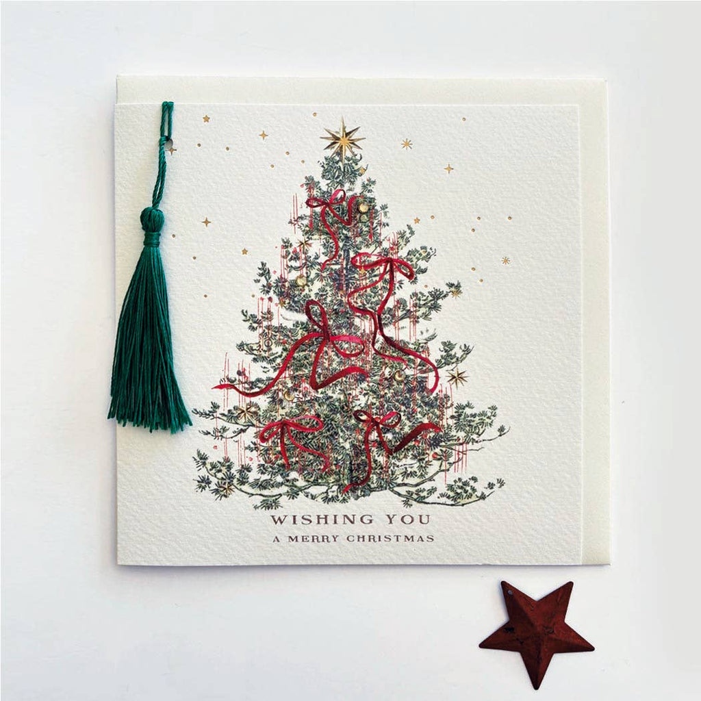 A square Christmas greeting card with a white textured background, featuring an illustrated decorated Christmas tree with red ribbons and a gold star. A dark green tassel hangs from the top-left corner, and the text 'WISHING YOU A MERRY CHRISTMAS' is printed at the bottom.