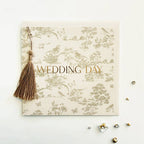 A square wedding keepsake album or card with a beige and gold toile de Jouy pattern, featuring the words 'WEDDING DAY' in gold foil. A brown tassel is attached to the left side, and the album is photographed on a white background with small decorative embellishments nearby.