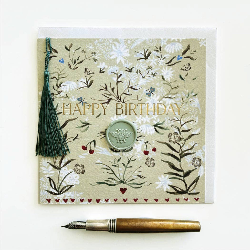 A square floral greeting card with the words “HAPPY BIRTHDAY” in gold lettering, decorated with botanical illustrations, a green tassel, and a round wax seal. A gold and brown fountain pen lies below the card on a clean white background.