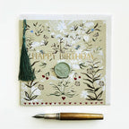 A square floral greeting card with the words “HAPPY BIRTHDAY” in gold lettering, decorated with botanical illustrations, a green tassel, and a round wax seal. A gold and brown fountain pen lies below the card on a clean white background.