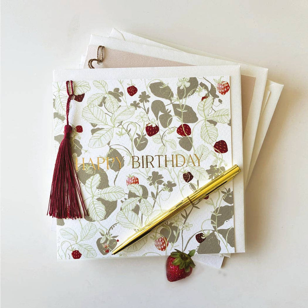 A square birthday card with a strawberry and leaf botanical pattern, gold 'HAPPY BIRTHDAY' text, a dark red tassel, and a gold pen resting on top, photographed on a clean white background.