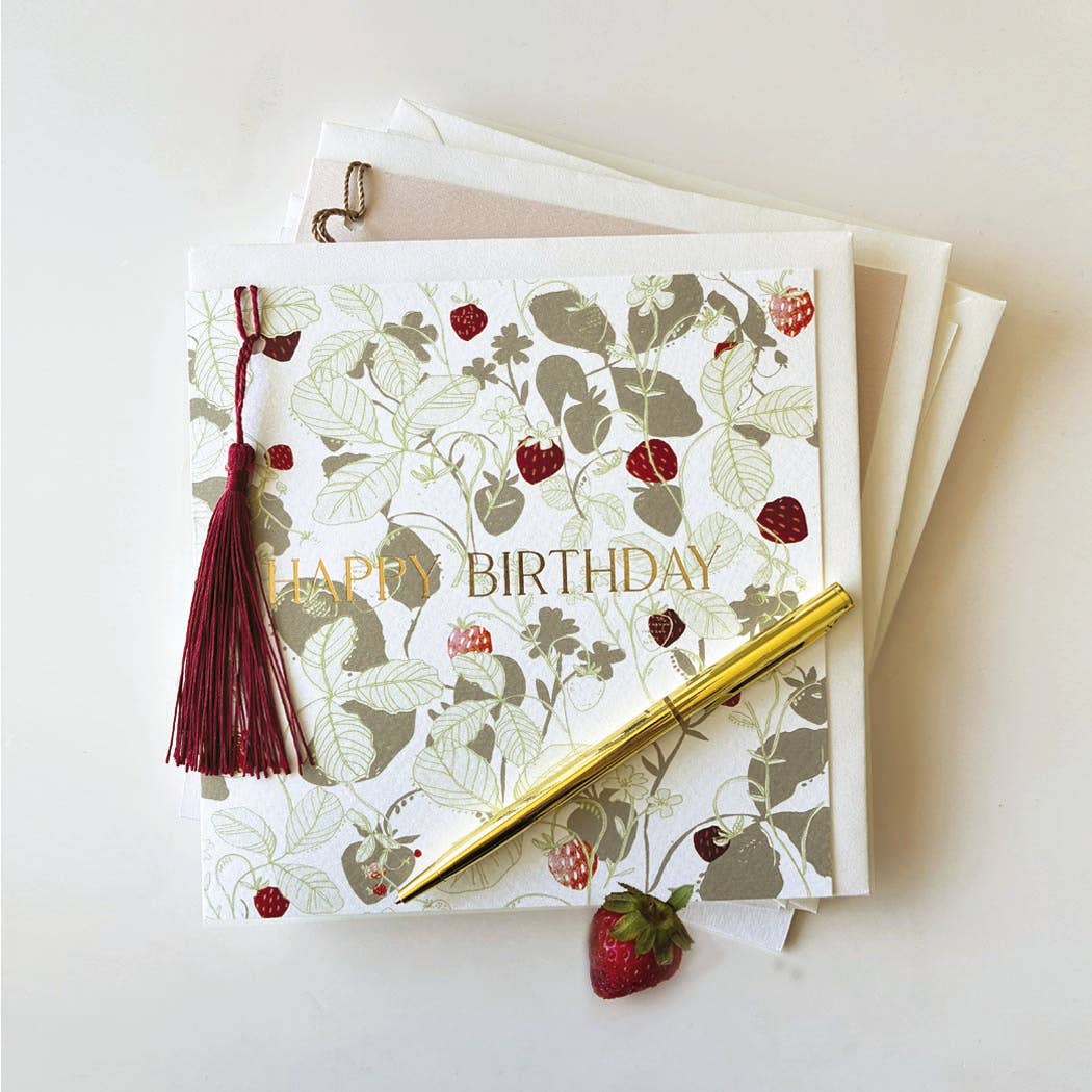 A square birthday card with a strawberry and leaf botanical pattern, gold 'HAPPY BIRTHDAY' text, a dark red tassel, and a gold pen resting on top, photographed on a clean white background.