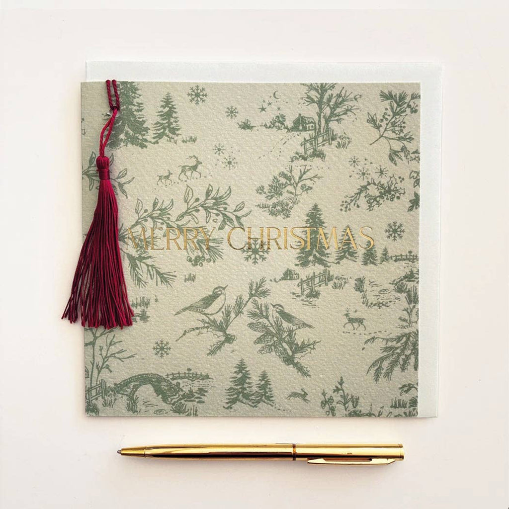 A square Christmas greeting card with a green toile de Jouy pattern of birds, trees, and snowflakes on a light beige background, featuring the words 'MERRY CHRISTMAS' in gold foil. A dark red tassel is attached to the left side, and a gold pen is placed below the card on a clean white background.