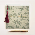 A square Christmas greeting card with a green toile de Jouy pattern of birds, trees, and snowflakes on a light beige background, featuring the words 'MERRY CHRISTMAS' in gold foil. A dark red tassel is attached to the left side, and a gold pen is placed below the card on a clean white background.
