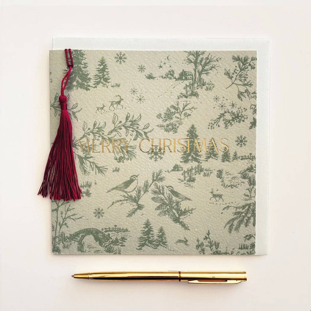 A square Christmas greeting card with a green toile de Jouy pattern of birds, trees, and snowflakes on a light beige background, featuring the words 'MERRY CHRISTMAS' in gold foil. A dark red tassel is attached to the left side, and a gold pen is placed below the card on a clean white background.